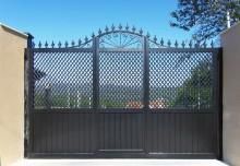 Puerta de metal negro con paneles de celosía, parte superior decorativa y listones inferiores verticales contra un cielo azul.