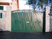 Puerta metálica verde entre pilares de piedra, situada en una zona pavimentada, con parte de un edificio visible.