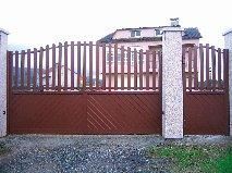 Puerta metálica de color marrón con lamas verticales sobre un panel macizo, delante de una casa.