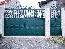 Puerta de metal verde con puerta peatonal a juego, herrajes ornamentados y pilares de hormigón.