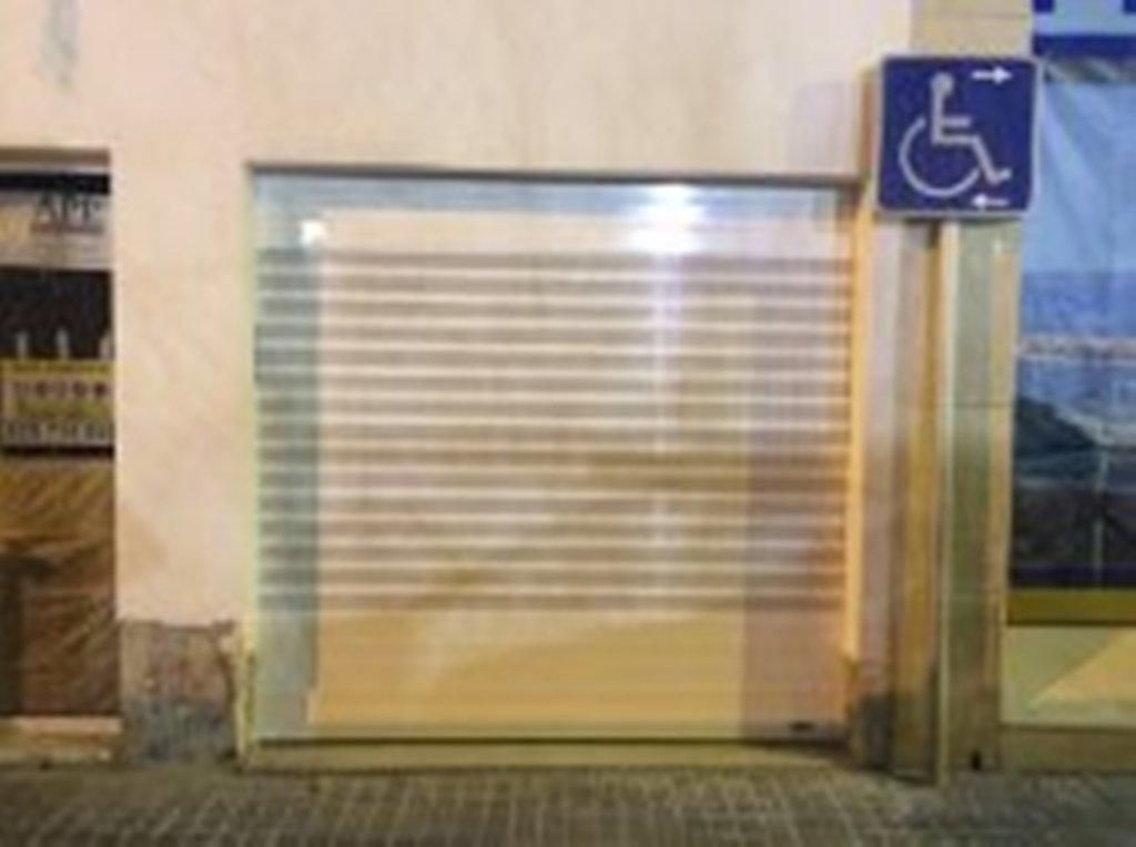 Una tienda cerrada con una persiana enrollable y una señal de aparcamiento para personas con discapacidad a la derecha. Paredes beige y acera de ladrillo.