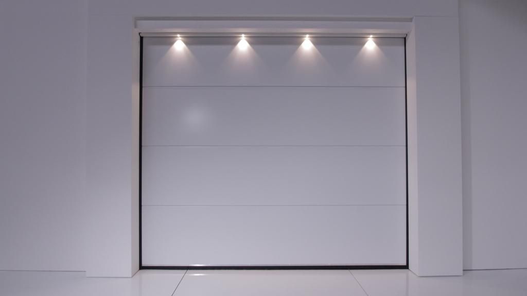 Puerta de garaje blanca, cerrada, con cuatro focos cenital, en un ambiente blanco y minimalista.