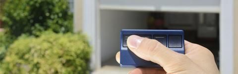 Botón presionado con la mano en el control remoto de la puerta del garaje azul frente a un garaje parcialmente abierto.