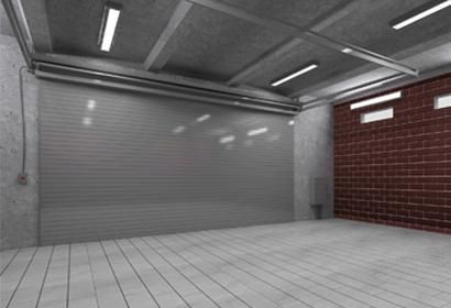 Interior de almacén vacío con puerta enrollable de metal cerrada, pared de ladrillo, piso y techo de concreto y luces fluorescentes.