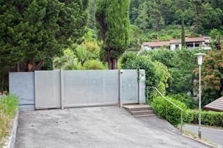 Una puerta de metal en un camino de entrada de hormigón conduce a una casa en una ladera con árboles al fondo.