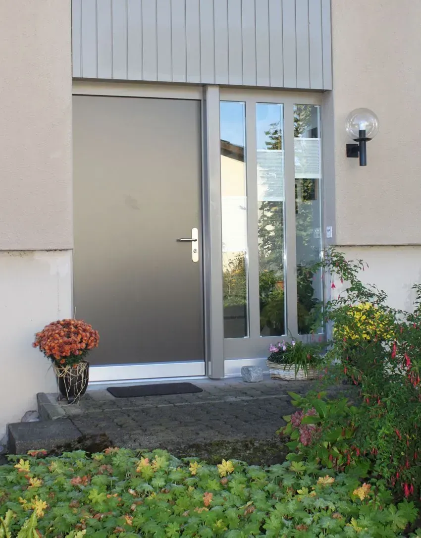 Vordereingang mit beiger Tür, seitlichen Glasfenstern, Wandleuchte und Blumenbeeten davor