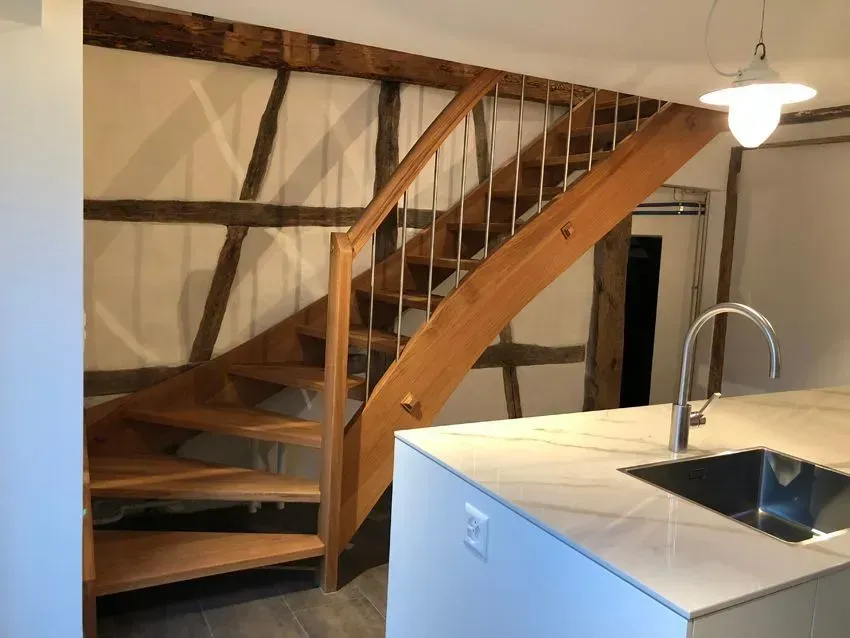 Eine Holztreppe mit offenen Stufen befindet sich in einem Raum neben einer Kücheninsel mit Spüle und moderner Pendelleuchte.