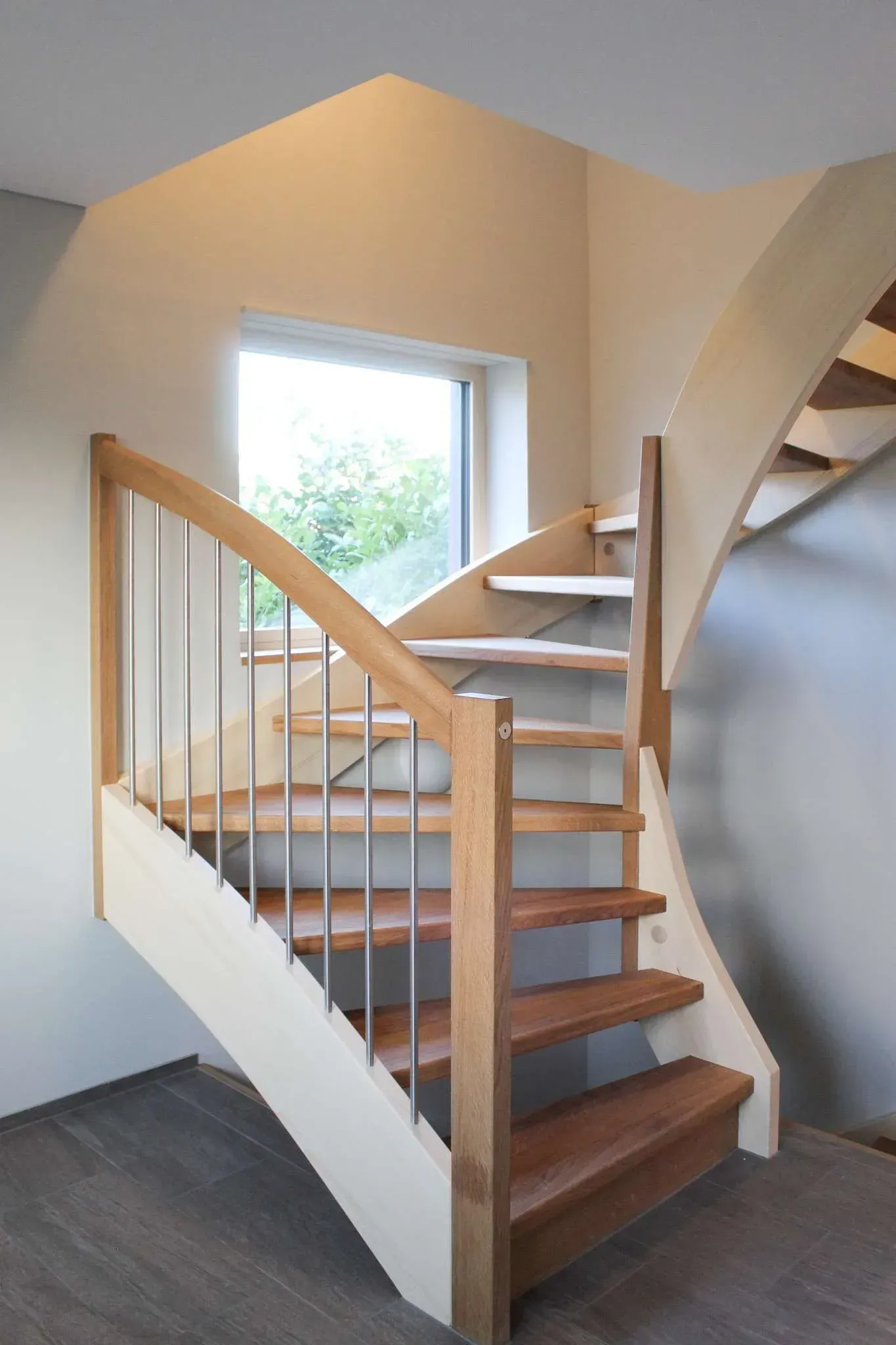 Holztreppe mit Geländer in einem hellen Innenraum, neben einem Fenster und einer geschwungenen Wand