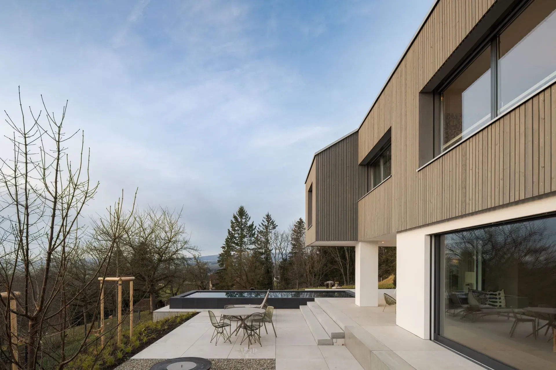 Modernes zweistöckiges Haus mit Holzverkleidung, großen Fenstern und einer gepflasterten Terrasse neben Garten und Pool
