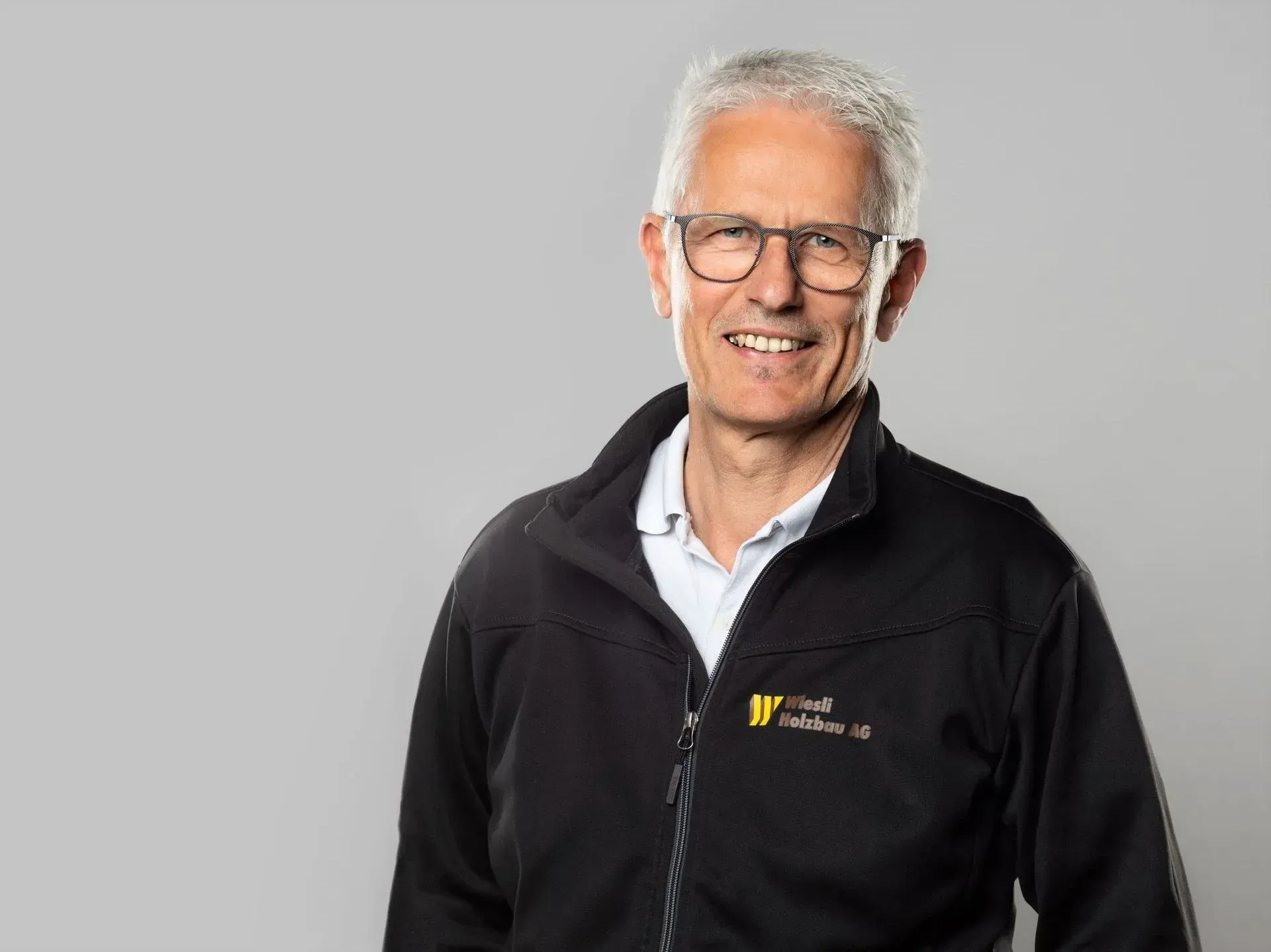 Lächelnder Mann mit Brille und schwarzer Jacke vor grauem Hintergrund