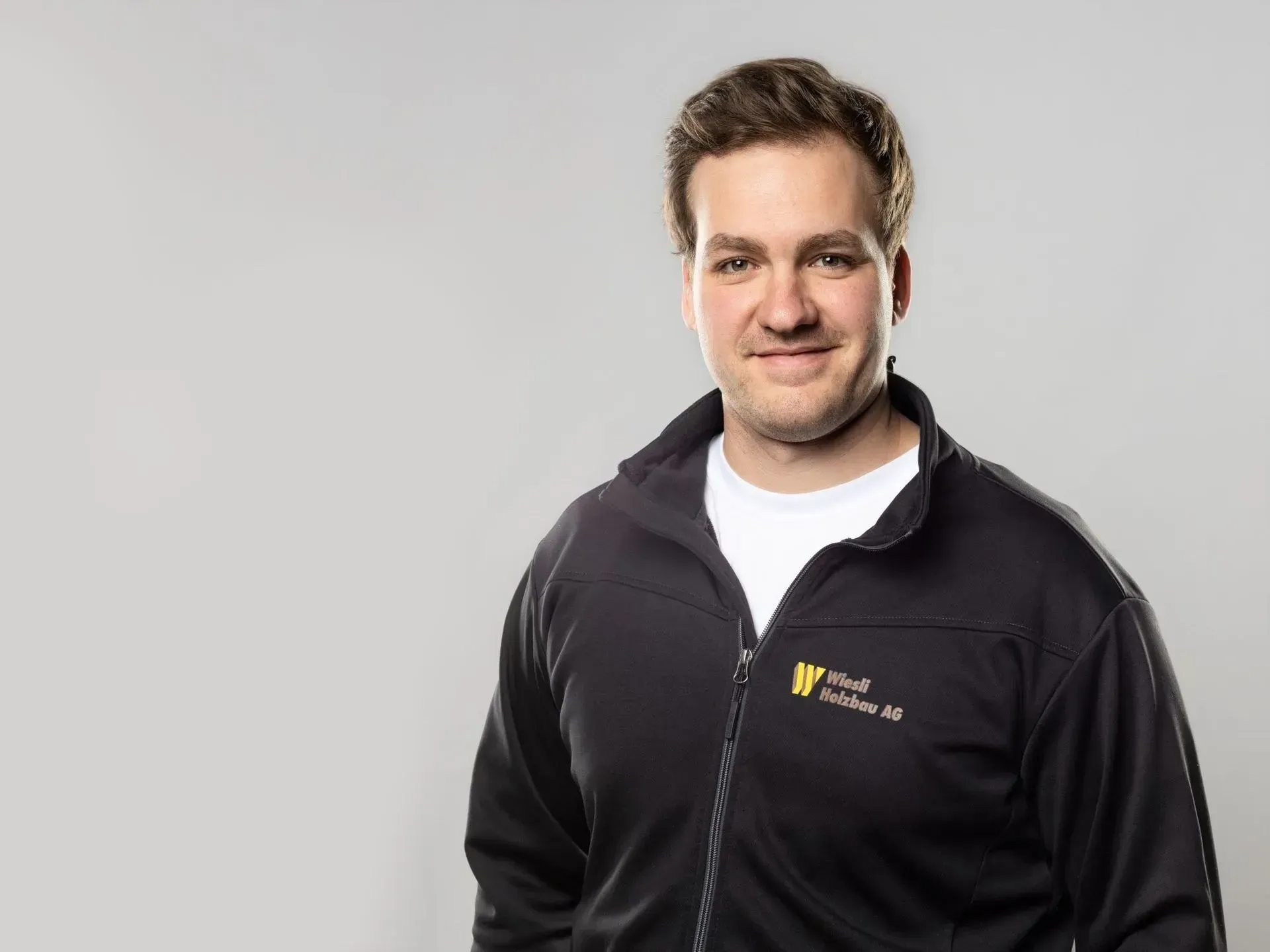 Mann in schwarzer Reißverschlussjacke mit Logo, steht vor hellgrauem Hintergrund und lächelt leicht