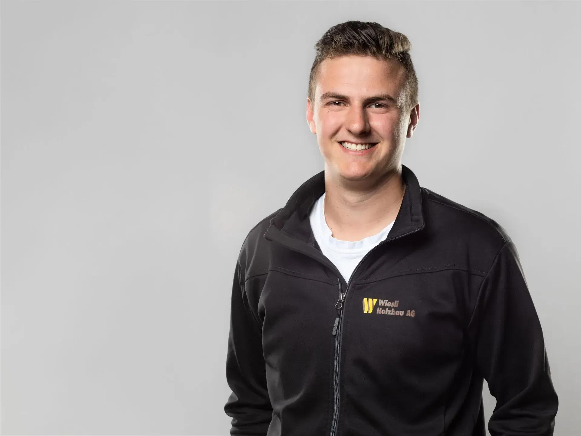 Lächelnder Mann in einer schwarzen Reißverschlussjacke mit Logo, vor hellgrauem Hintergrund