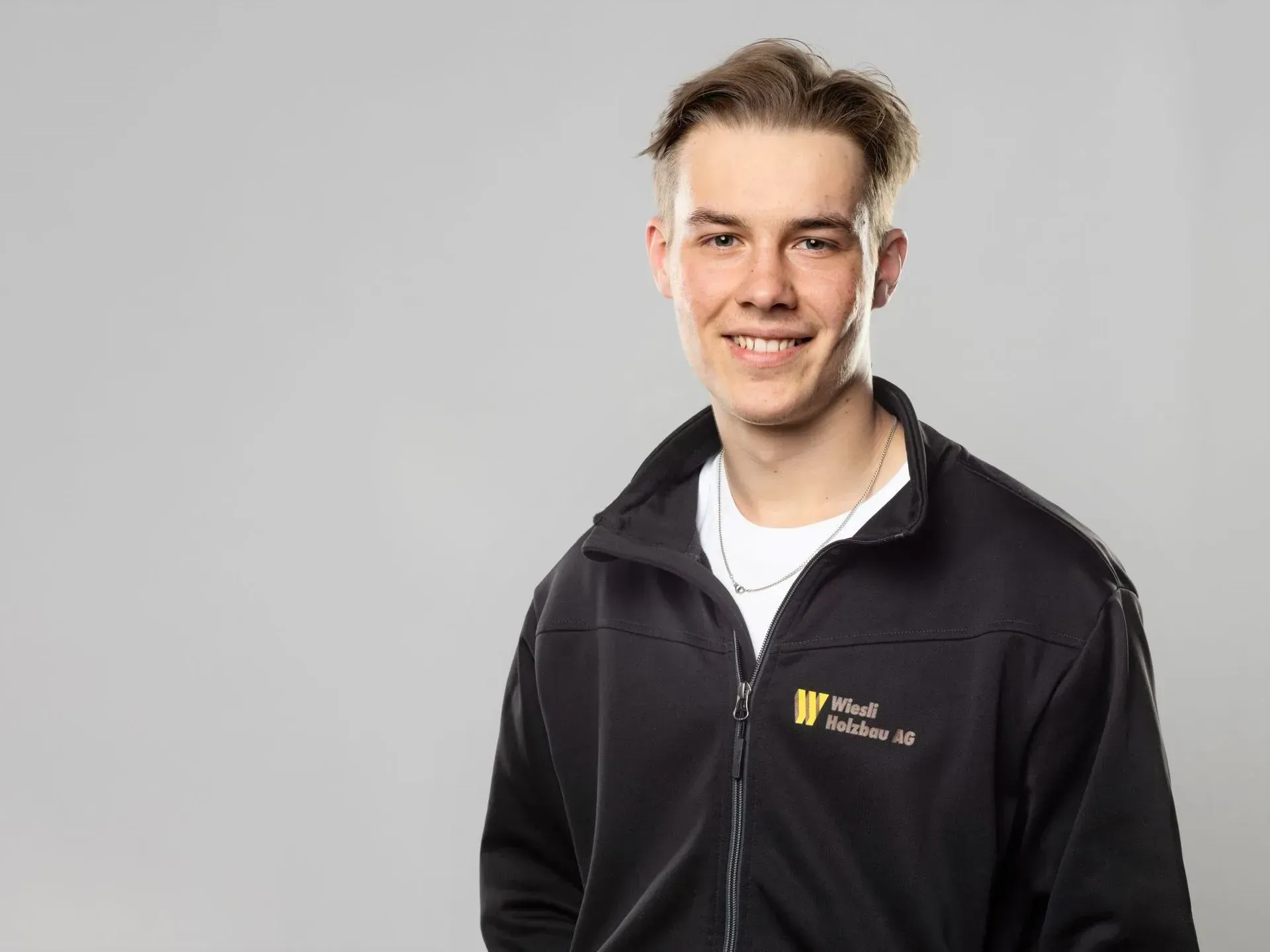 Lächelnder Mann in einer schwarzen Reißverschlussjacke mit Logo, vor hellgrauem Hintergrund