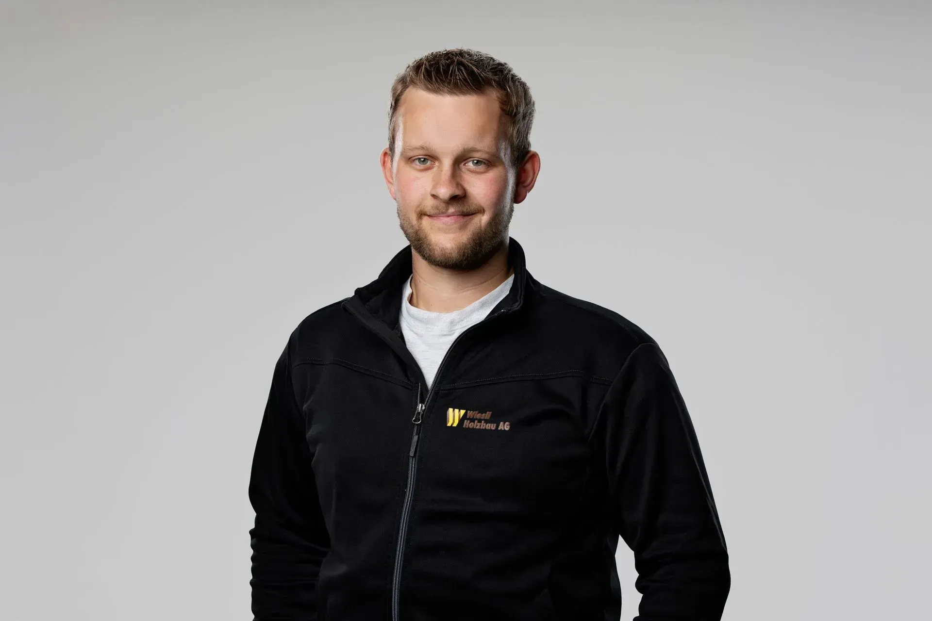 Mann in einer schwarzen Reißverschlussjacke, der vor einem hellgrauen Hintergrund steht und in die Kamera blickt.