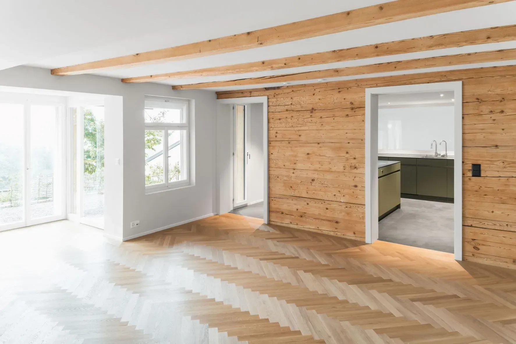 Heller, leerer Raum mit freiliegenden Holzbalken, hellen Fußböden und einem Durchgang zur Küche.