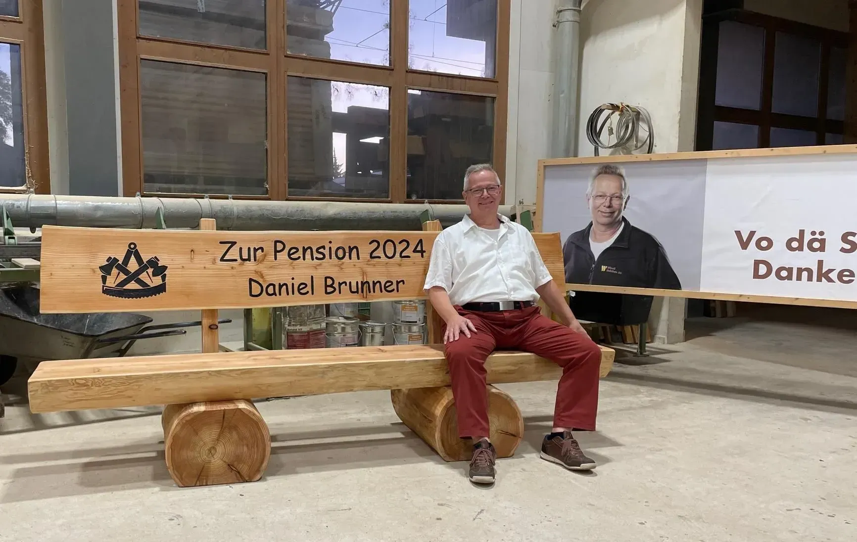 Ein Mann sitzt auf einer Holzbank in einem rustikalen Zimmer neben einem Schild mit der Aufschrift „Zur Pension 2024 Daniel Brugner“.