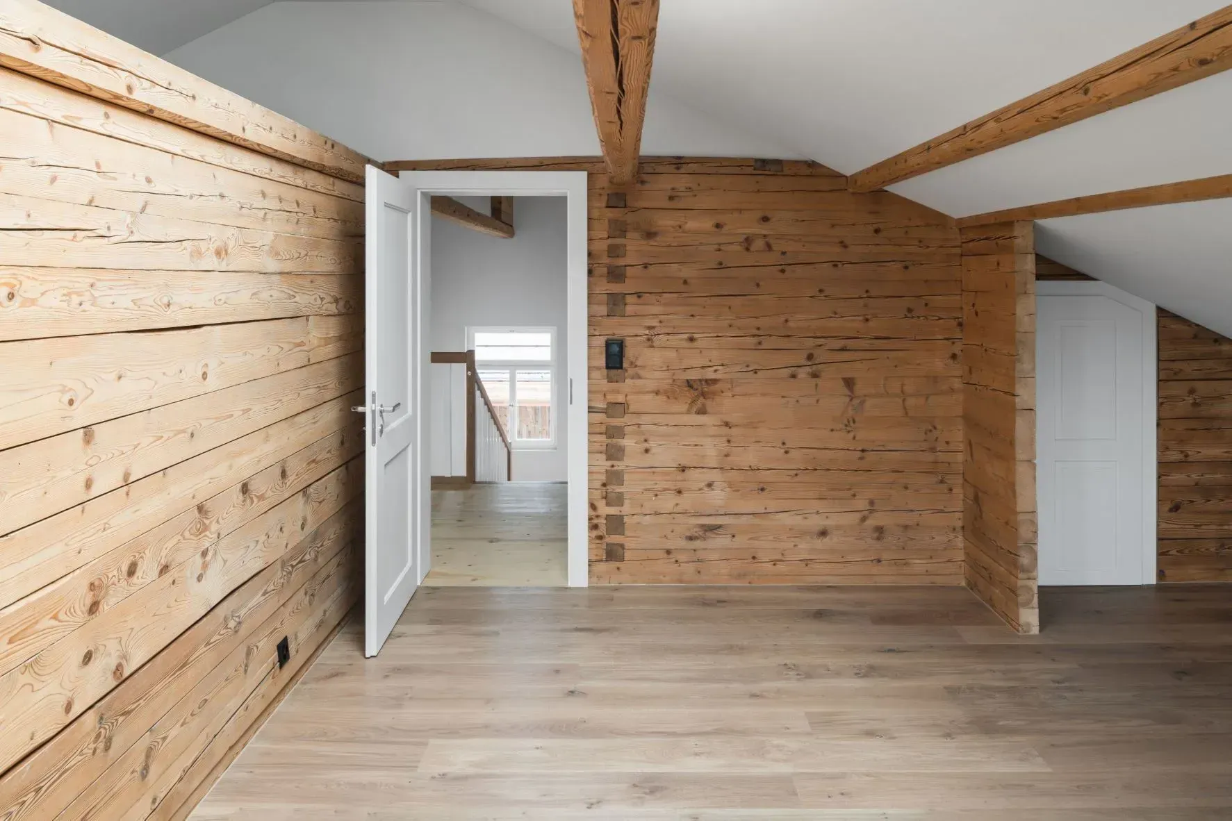 Leeres Dachzimmer mit hellen Holzwänden, weißer Tür und sichtbaren Balken.