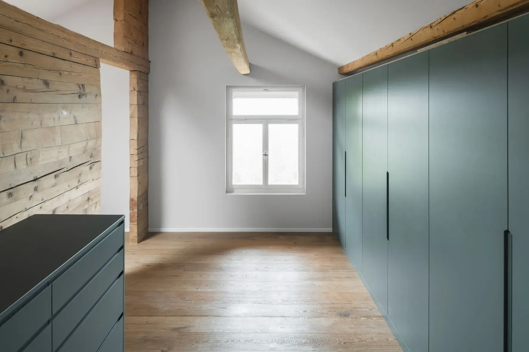 Helles, leeres Zimmer mit Holzboden, türkisfarbenen Schränken, sichtbaren Balken und einem Fenster.