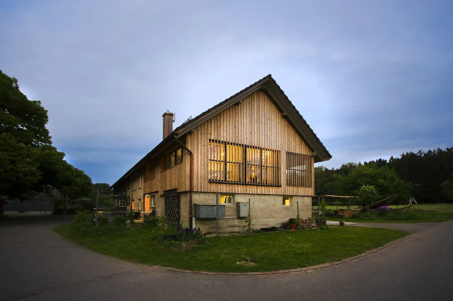 Holzhaus in der Abenddämmerung mit warm erleuchteten Fenstern, umgeben von Gras und Bäumen unter einem bewölkten Himmel