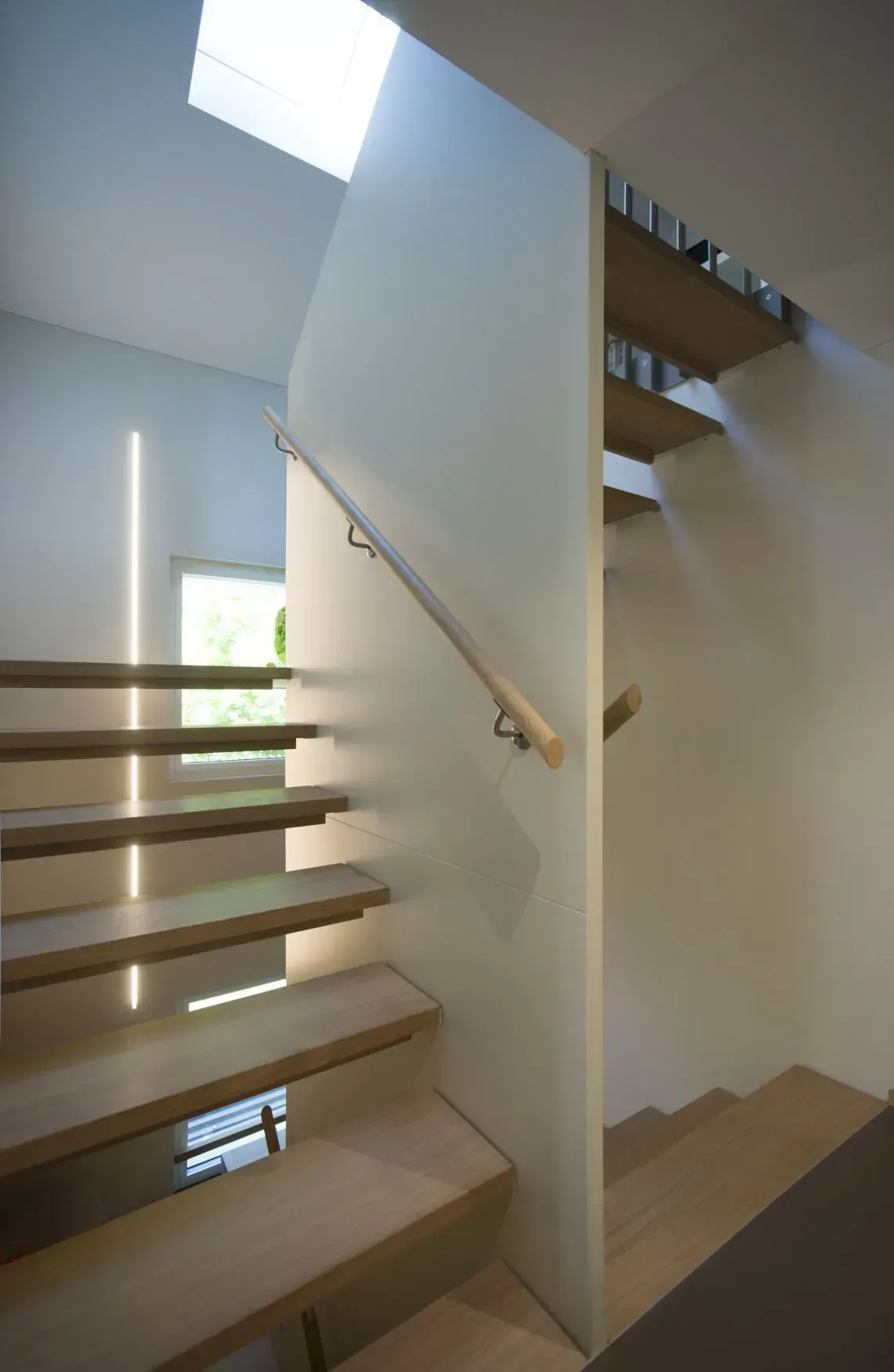 Moderne Innentreppe mit Holzstufen, weißen Wänden und einem Handlauf neben einem lichtdurchfluteten Treppenabsatz