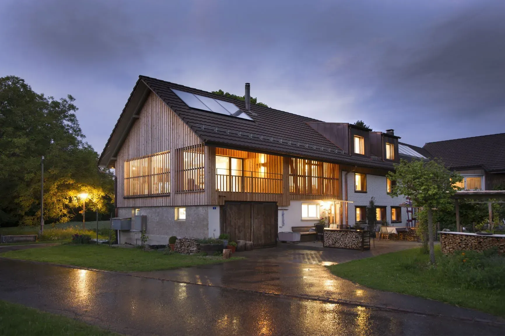 Modernes Haus mit warm beleuchteten Fenstern in der Abenddämmerung, die sich in einer nassen Straße spiegeln.