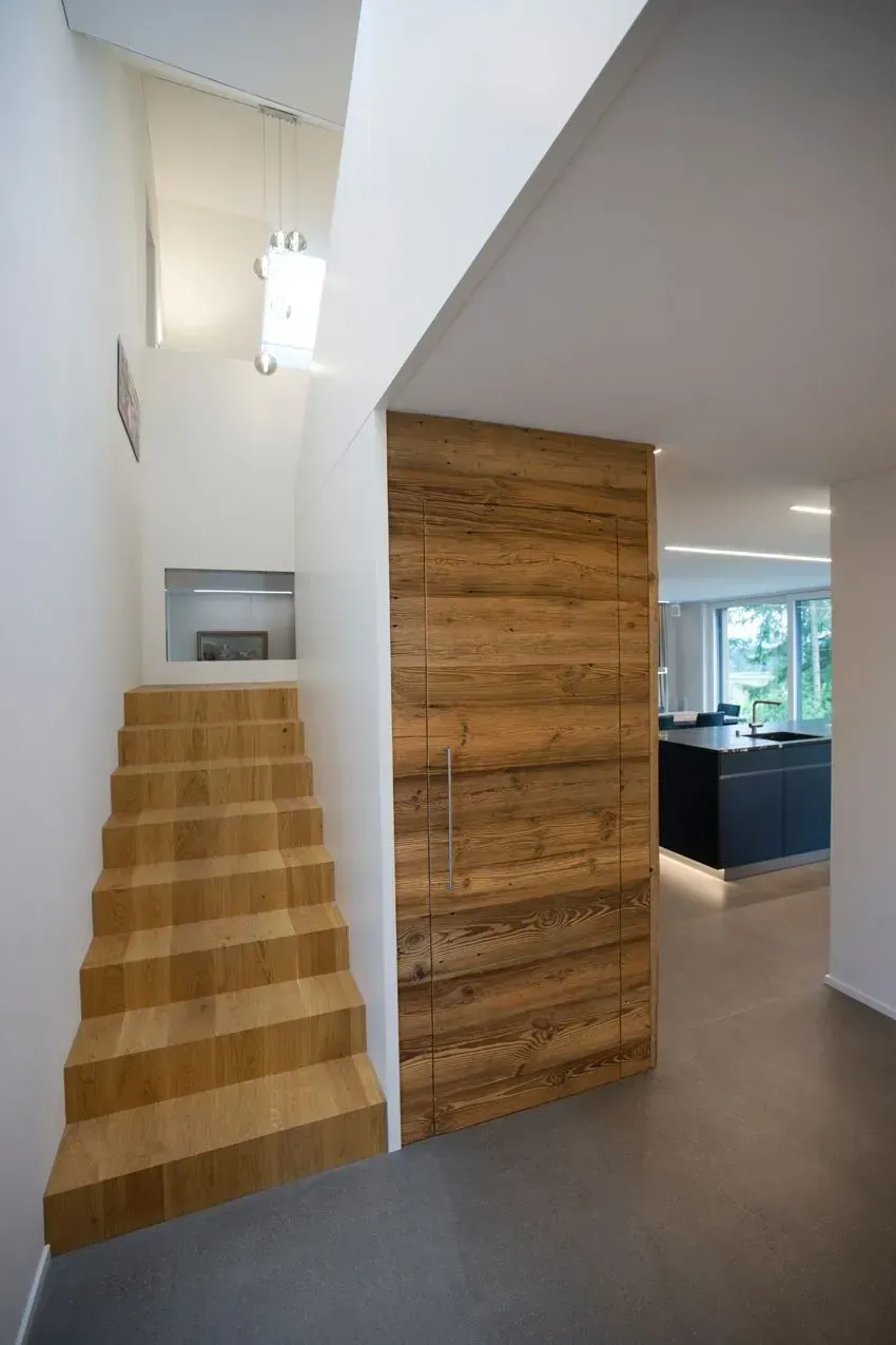 Modernes Interieur mit holzverkleideter Treppe, weißen Wänden, grauem Boden und einer im Hintergrund sichtbaren Küche.