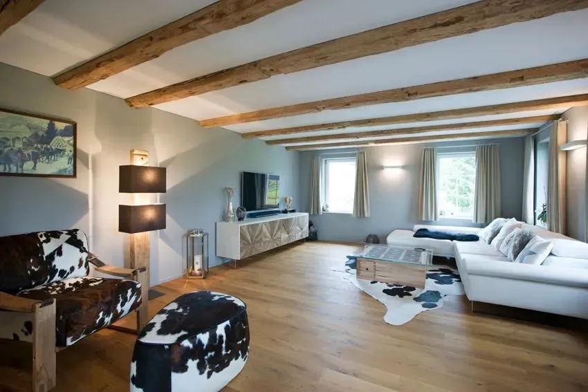 Helles Wohnzimmer mit weißer Eckcouch, Holzbalken und großen Fenstern mit Blick auf die Bäume