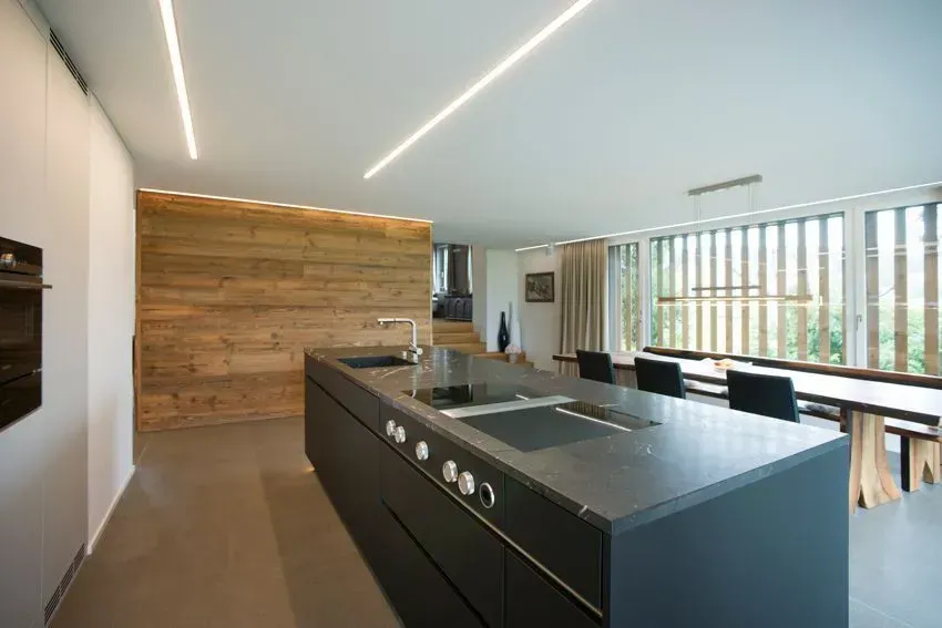 Moderne offene Küche mit dunkler Kochinsel, Akzentwand aus Holz und großen Fenstern mit Blick ins Grüne