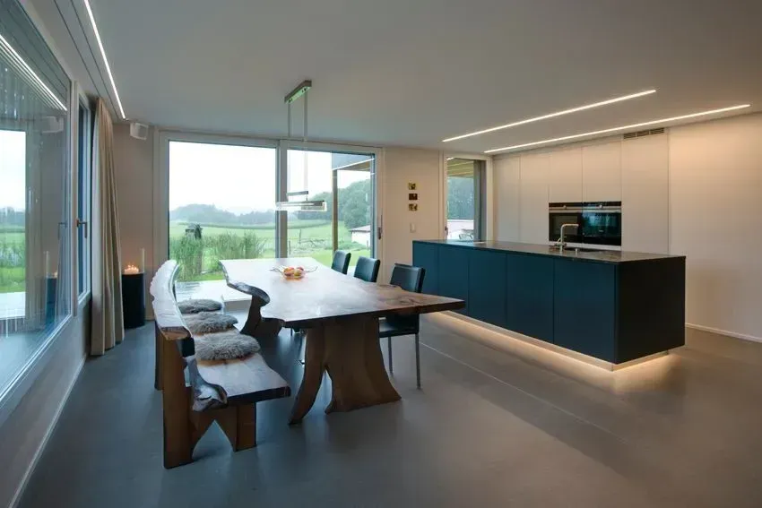 Modernes Esszimmer mit langem Tisch, weißen Stühlen, dunkelblauer Kücheninsel und großen Fenstern mit Blick auf die Landschaft