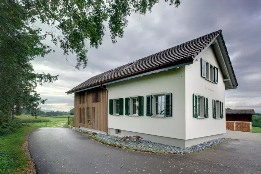 Weißes Haus mit grünen Fensterläden auf einer Kiesauffahrt, umgeben von Bäumen in einer ländlichen Landschaft