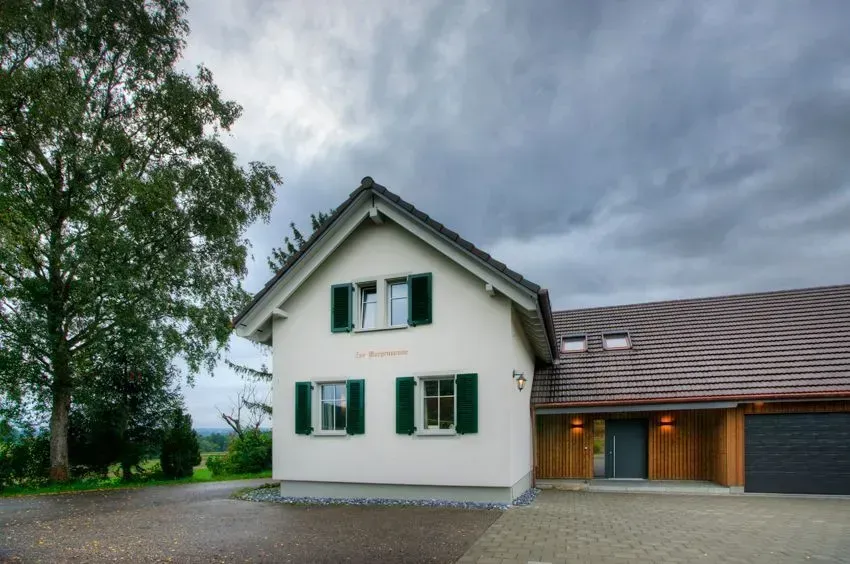 Weißes Haus mit grünen Fensterläden und dunklem Dach neben einer Garage unter bewölktem Himmel