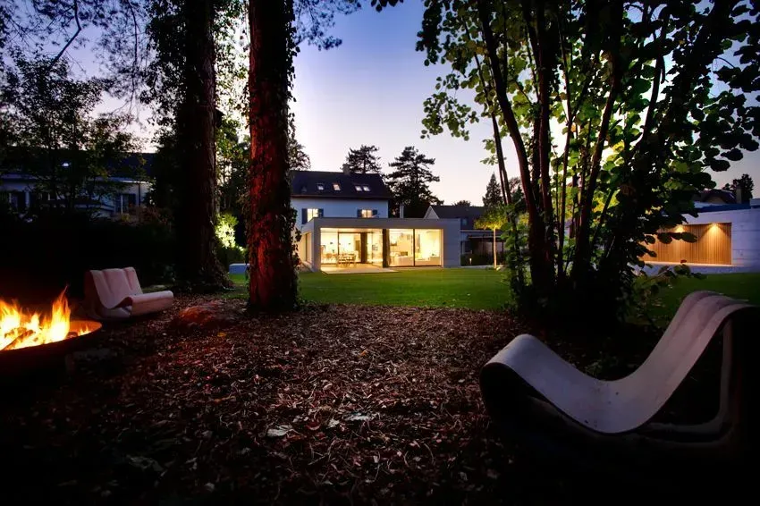Ein im Dämmerlicht erstrahlender Hinterhof mit Liegestühlen, Feuerstelle und einem modernen Haus, das im Hintergrund leuchtet.