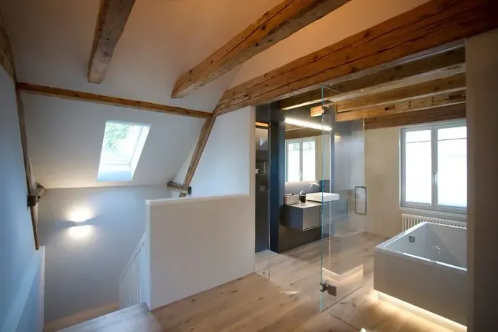 Helles Dachgeschossbadezimmer mit freiliegenden Holzbalken, Oberlicht und Glasdusche neben einer Badewanne.
