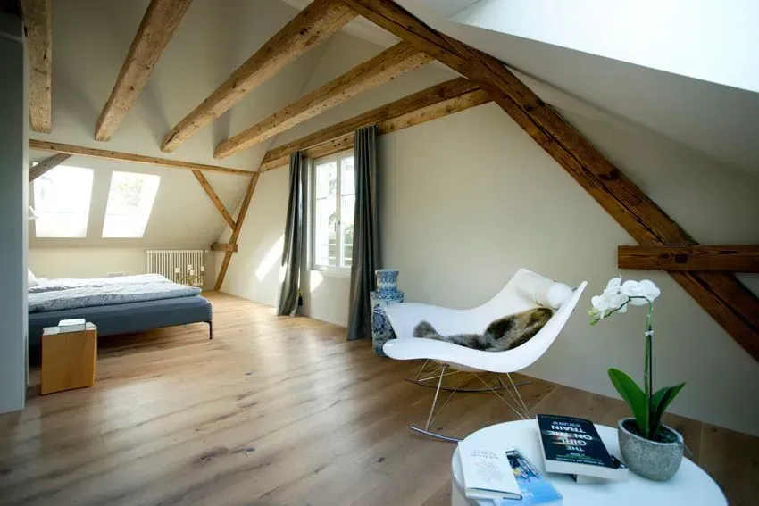 Helles Dachgeschoss-Schlafzimmer mit sichtbaren Balken, Dachfenstern, einem Bett, einem weißen Sessel und Holzböden