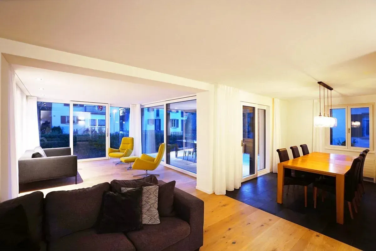 Modernes Wohn-Esszimmer mit dunklem Sofa, gelben Stühlen, Holztisch und großen Fenstern mit Blick auf die Abendlichter
