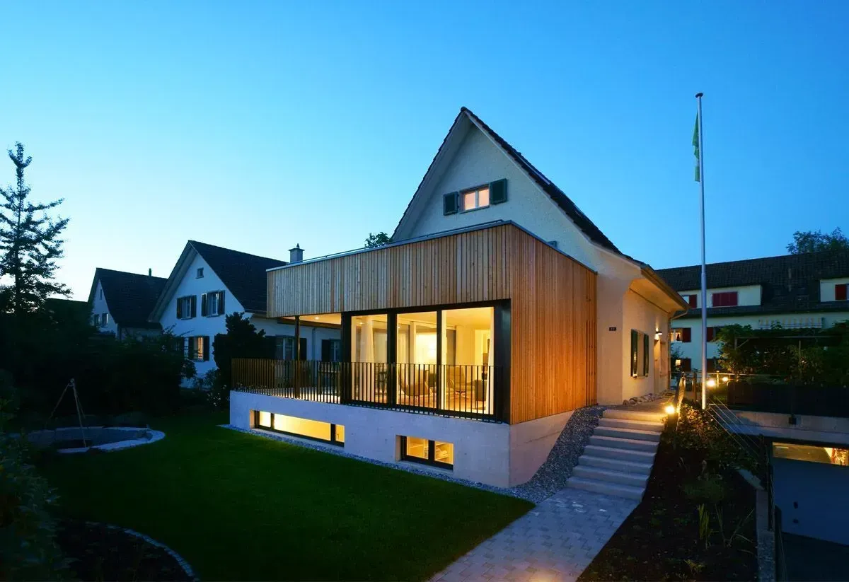 Modernes Haus mit warm beleuchteten Fenstern in der Abenddämmerung, umgeben von einem Rasen und Nachbarhäusern.