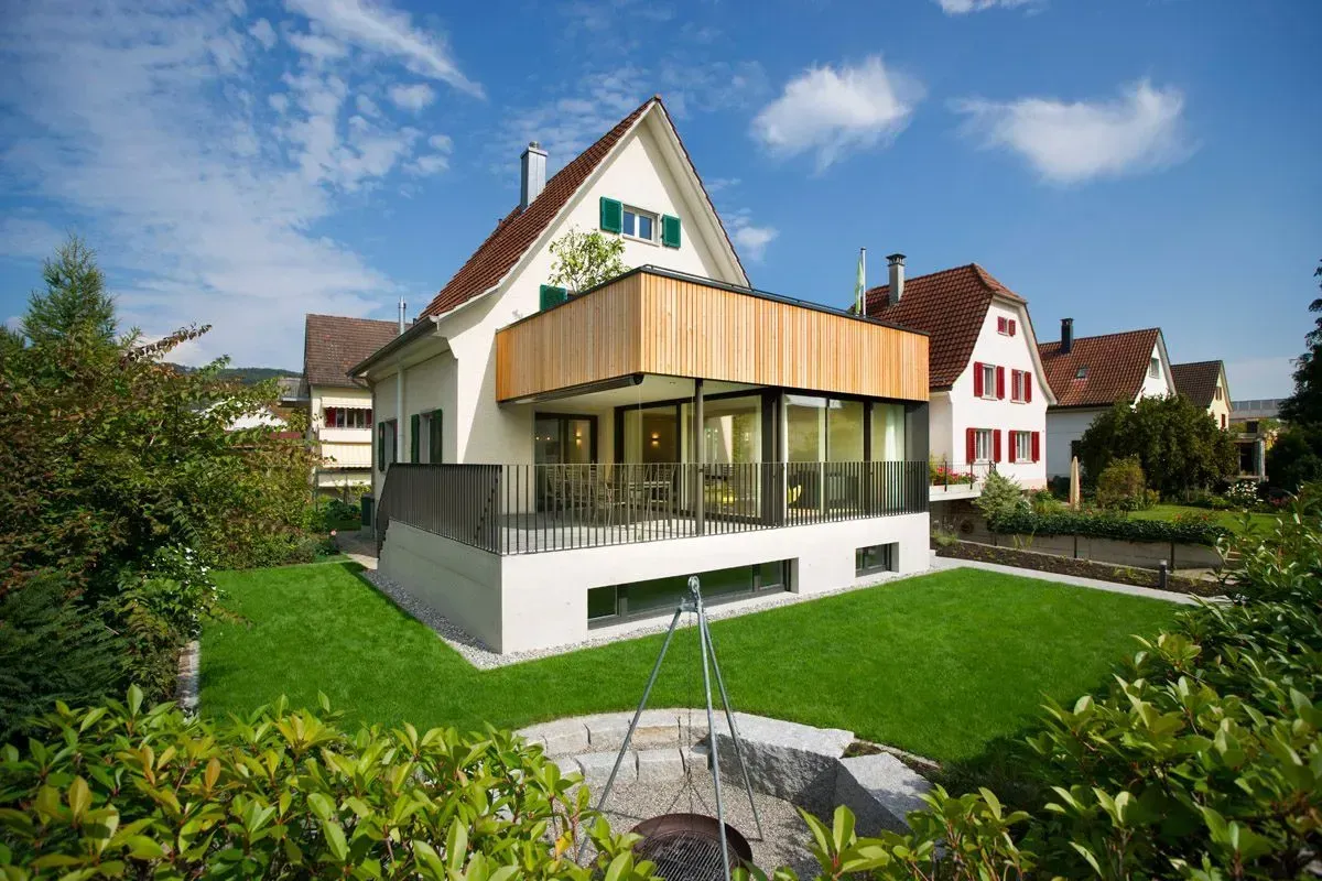 Modernes weißes Haus mit Holzelementen, umgeben von grünem Rasen und einem kleinen Brunnen unter blauem Himmel