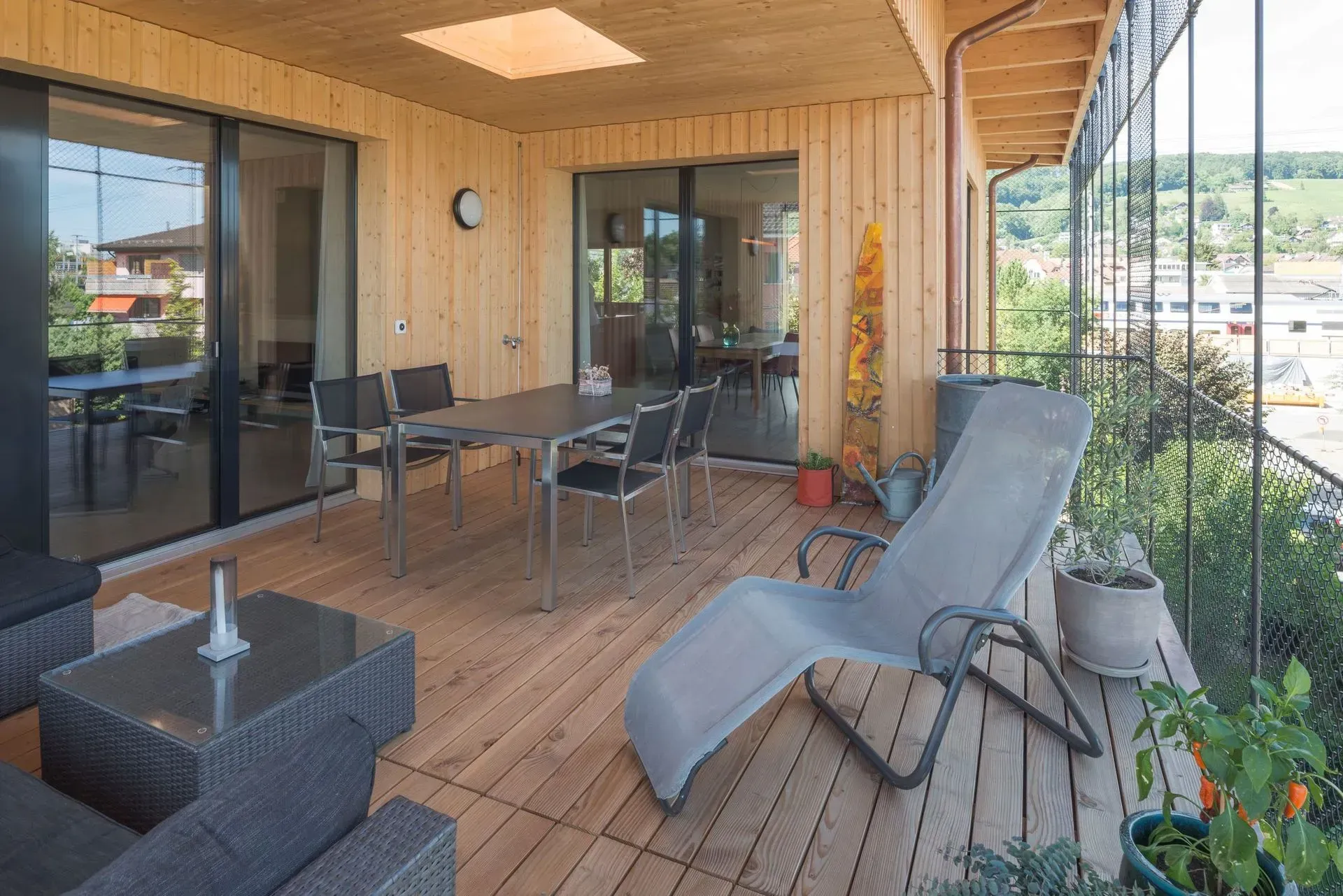 Überdachte Terrasse mit Korbmöbeln, Esstisch und Liegestuhl neben Glasschiebetüren.