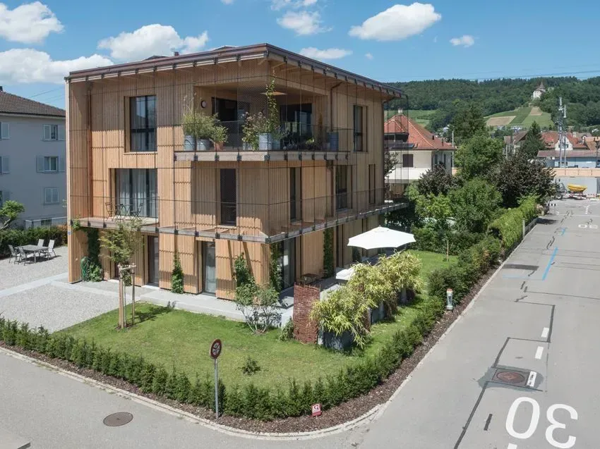 Modernes Holzhaus auf einem Eckgrundstück mit Balkonen, Grünflächen und einer ruhigen Straße unter blauem Himmel