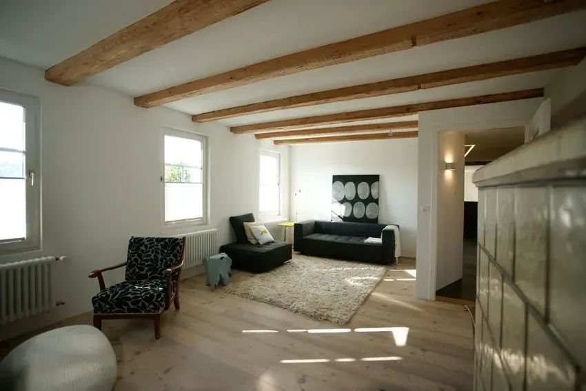 Helles Wohnzimmer mit sichtbaren Holzbalken, Sofa, Sessel, Teppich und Sonnenlicht durch die Fenster