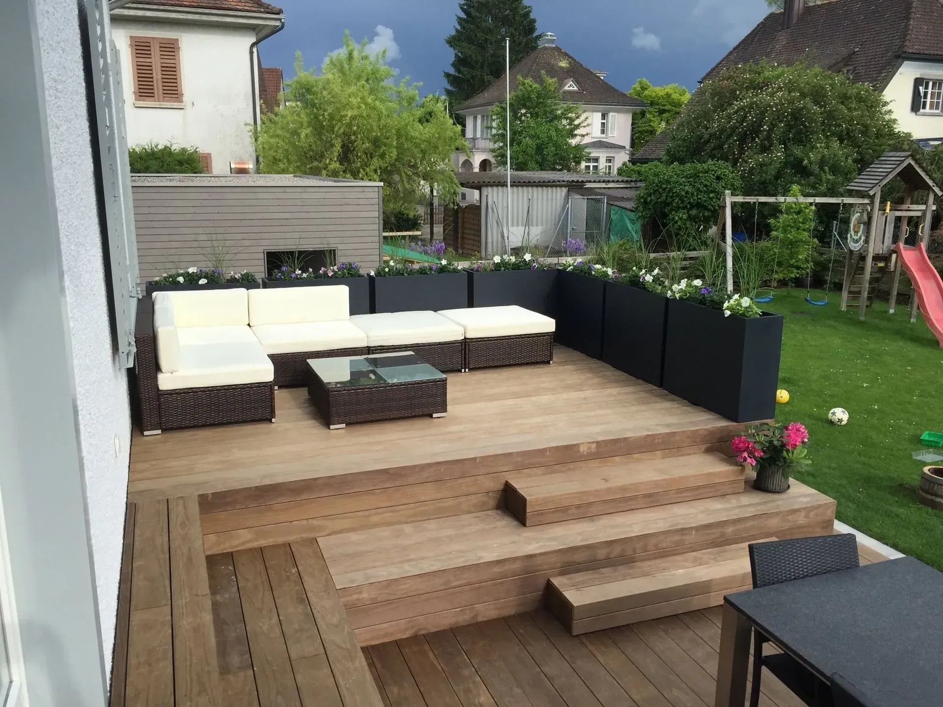Holzterrasse im Garten mit weißem Gartensofa, schwarzem Hocker, Treppe und Rasen mit Schaukel im Hintergrund