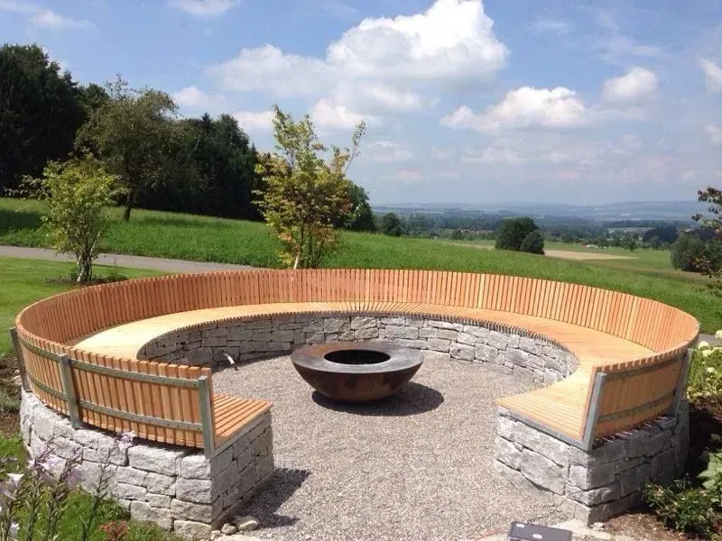 Runde Feuerstelle im Freien mit Holzbank, Steinsockel und Blick auf die Landschaft unter einem teilweise bewölkten Himmel