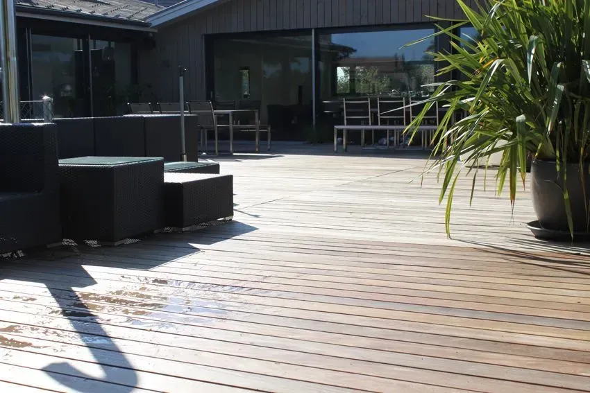 Sonnendurchflutete Holzterrasse mit dunklen Sitzgelegenheiten im Freien und einem modernen Gebäude mit Glastüren