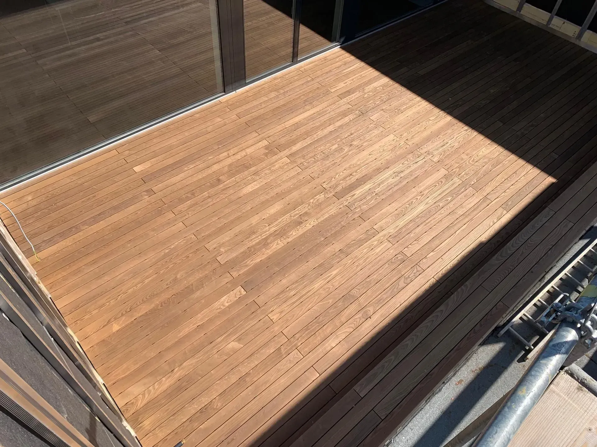Holzterrasse mit sonnigen und schattigen Bereichen neben einem Geländer und Metallrohren