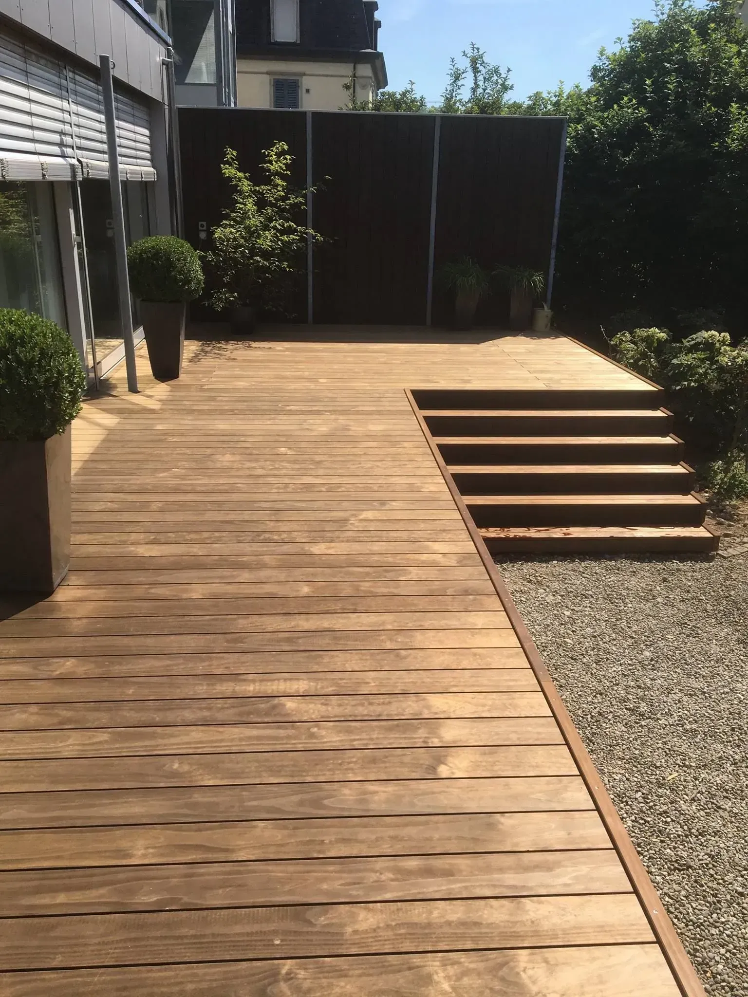 Ein hölzerner Terrassenweg mit Stufen neben einem Kiesweg führt zu einem umzäunten Hinterhof im hellen Sonnenlicht.