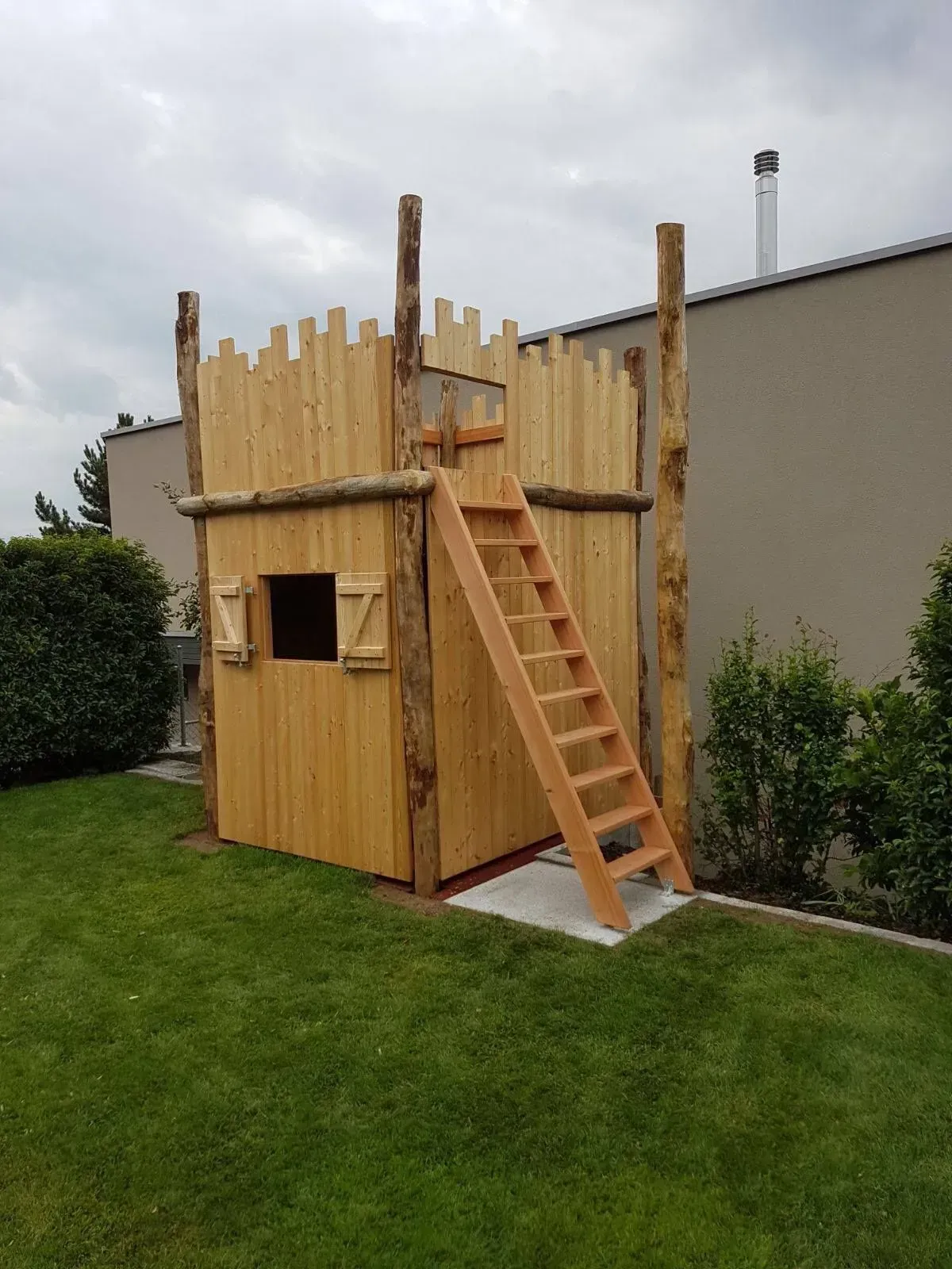 Spielhaus aus Holz mit Leiter in einem grasbewachsenen Hof neben einem Haus