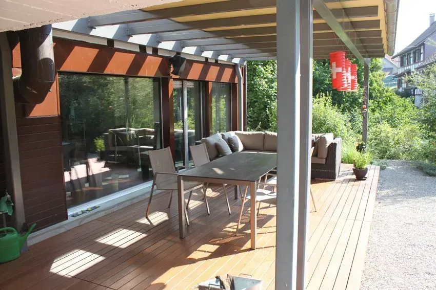 Überdachte Holzterrasse mit Esstisch, Stühlen, Grill und Gartenblick neben einem Kiesweg