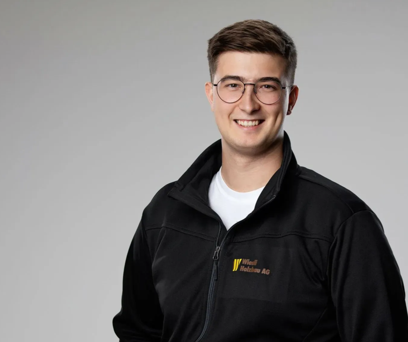 Person mit Brille und schwarzer Jacke lächelt vor einem einfarbigen grauen Hintergrund