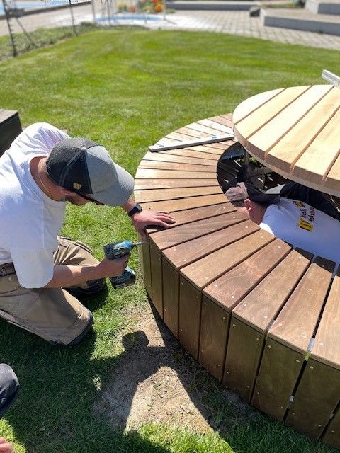Eine Person kniet neben einer Holzbank und telefoniert in einem sonnigen, grasbewachsenen Parkbereich.
