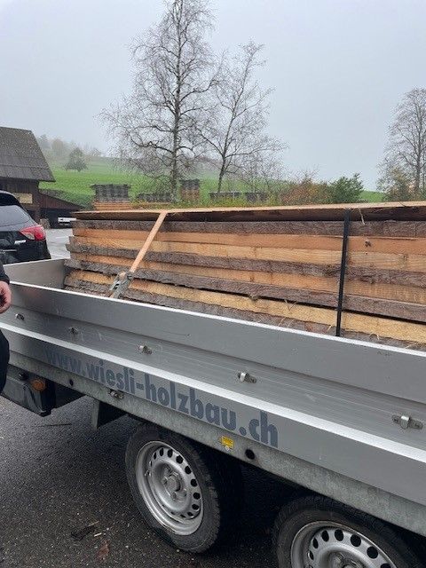 Grauer Pritschenanhänger, beladen mit gestapelten Holzbrettern und einem Metallgeländer im Freien